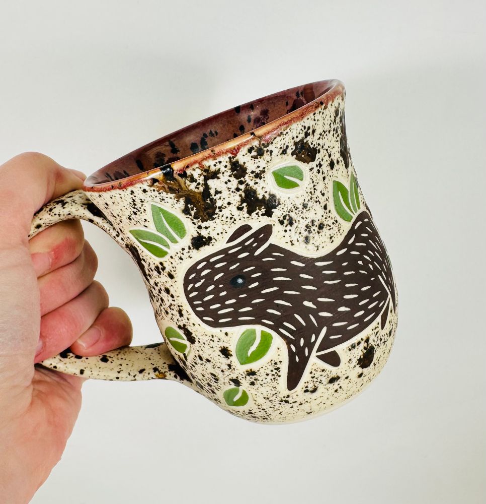 A handmade mug with a capybara illustration on it. The mug is glazed in a brown speckle glaze on the outside and a reddish brown glaze on the inside.