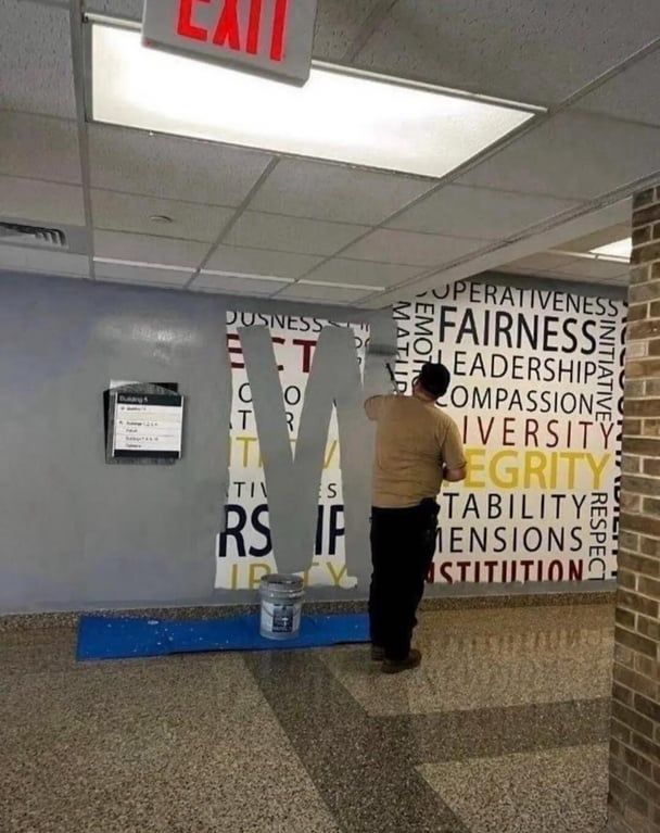 A photograph hallway with large brightly colored words including "Fairness, integrity, compassion, diversity, leadership, cooperstiveness, and respect" is half covered by prison cell gray paint that a man is applying with a paint roller 