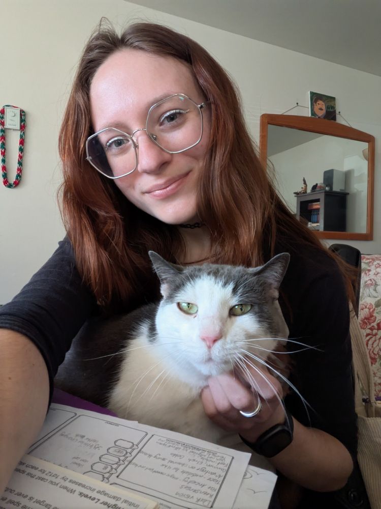Selfie of Jess with a grey and white cat on her lap. Jess has auburn hair, silver hexagonal glasses, and white winged eyeliner. The cat has green eyes and a pink nose and is receiving a scritch under the chin.