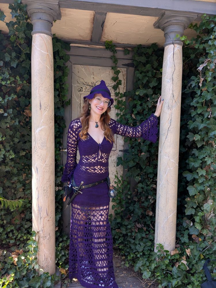 Jess posing between two marble-painted columns with ivy growing on the walls around them. The dress she's wearing is an eggplant purple crocheted masterpiece with a netting pattern on the long bell sleeves and full skirt. There's a matching purple witchy hat with a floppy brim and black ribbon around. A belt is slung on her hips with a dagger on the left and a round potion bottle on the right.