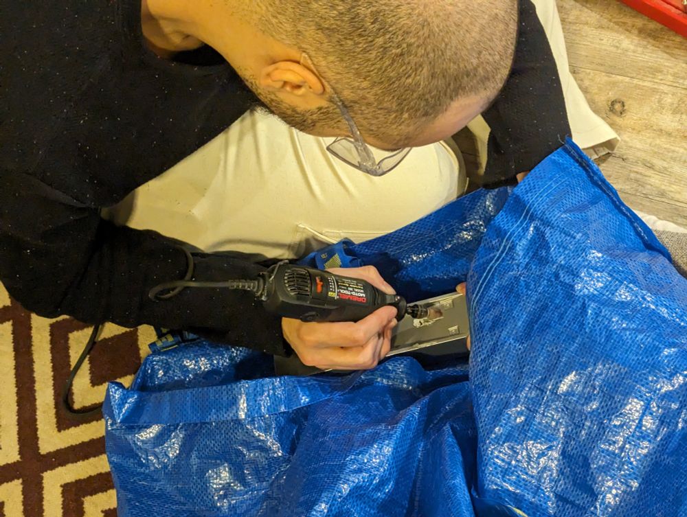top view of a nearly bald man wearing clear safety glasses and using a hand-held Dremel to cut into a grey plastic rectangle being held above a large blue ikea ahopping nag