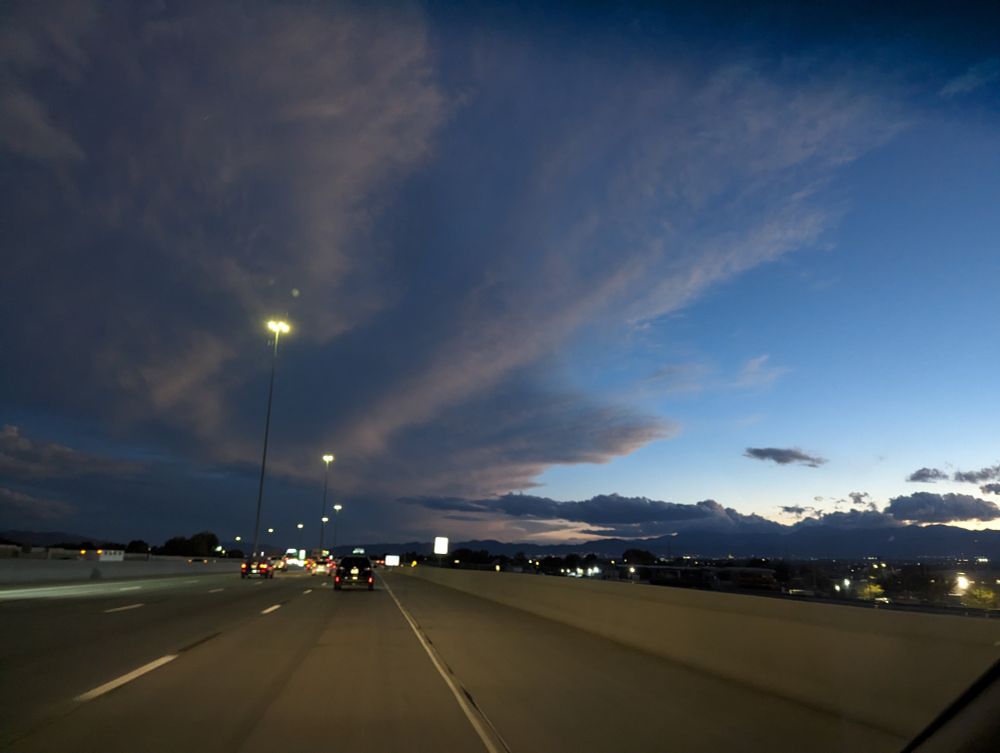 pov from car on freeway. Large grey clouds roll from the left to the right, stopping a out halfway. Blue skies on the right. Street lamps on the left of the road snake into the distance.  