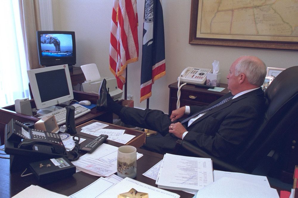 An older man in a dark suit sits at a desk in an office with his leg raised on the desk, watching a small television screen that shows the Twin Towers burning. The desk has multiple telephones, papers, and a coffee mug. It’s Dick Cheney on September 11, 2001. 