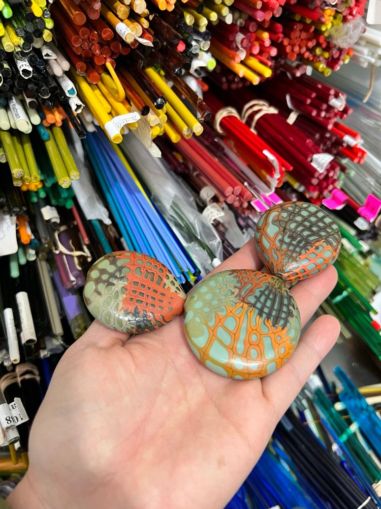 Hand blown and tumble etched glass beads in a flat tab shape. The grid stringer work recalls an old design redone off mandrel. Reminiscent of fish nets or darning, eventually everything frays and needs repair. The base color is a sort of mineral green that reminds me of cactus and the two flat drop shaped beads on either side evoke cactus paddle shapes. 
I’ve been building up inventory for Glasscraft Bead Expo this coming April 2-6th in Las Vegas, my first time back since 2019! Give a shout out if you’ll be there, too- it’ll be great to meet more glass art people. 
The background shows the glass rods that get melted to eventually become beads. 🔥
#ajavazglass #offmandrelbeads #blownbeads #flameworking #lampworking #tumbleetchedbeads 