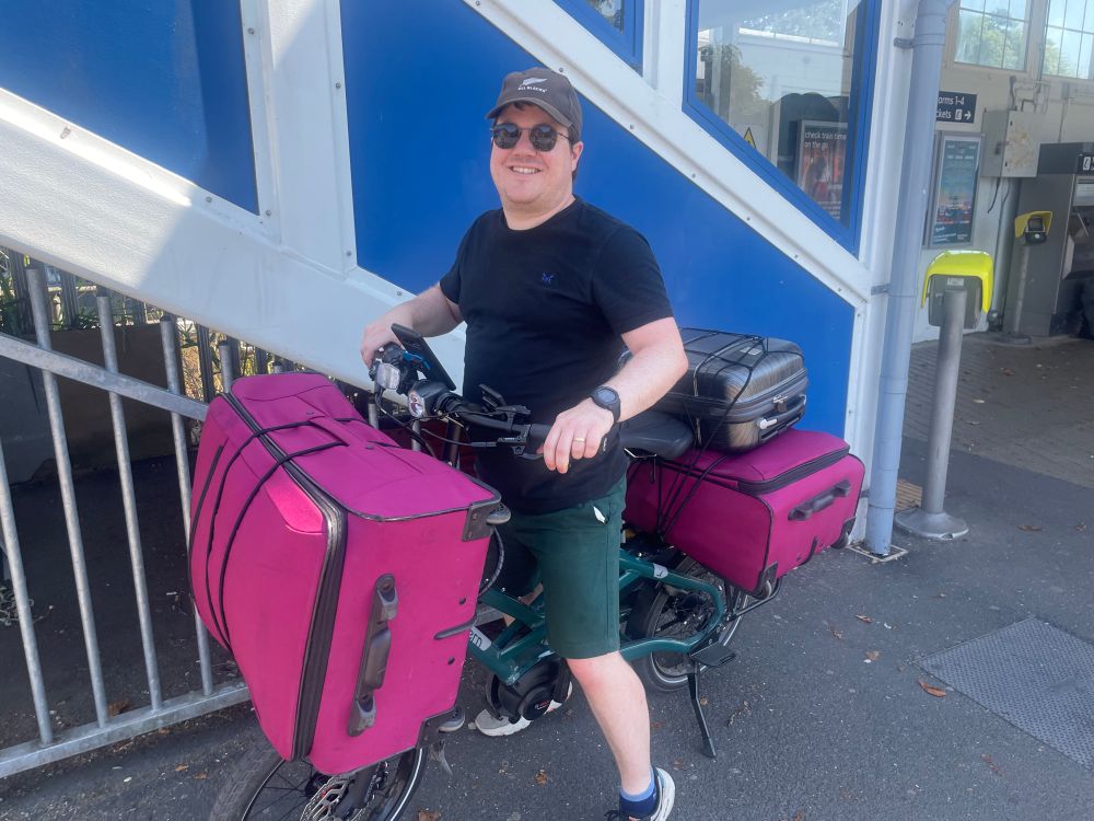 Photo shows a man on a green bike outside a railway station. There is a large pink suitcase strapped to the front of the bike, and a large pink suitcase strapped to the rear rack, with a small black case on top.
