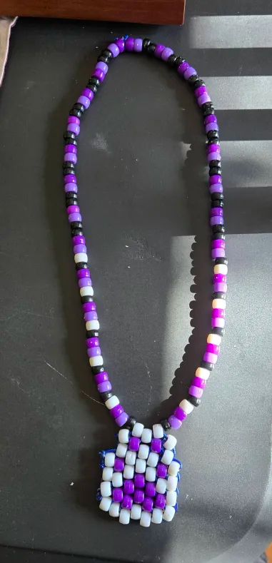 A pony bead necklace. The necklace part is black, white, light purple, and dark purple. The charm is also made from pony beads and it is a white and purple paw, with the paw being purple and the outside edges being white.