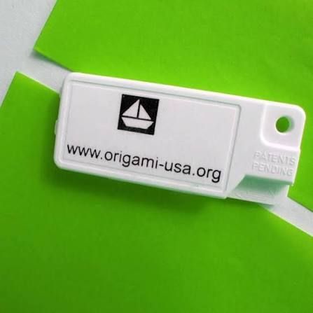 a white envelope opener, called the "envelopener". it's a white rectangular piece of plastic that guides a folded piece of paper to perfectly cut along the crease with a captive blade (perfect for cutting paper for folding origami on the go, no bulky guillotime paper cutter required!) this one is branded with a black and white origamiusa logo (a geometric sailboat) and has "www.origami-usa.org" printed below on the side of the cutter. 