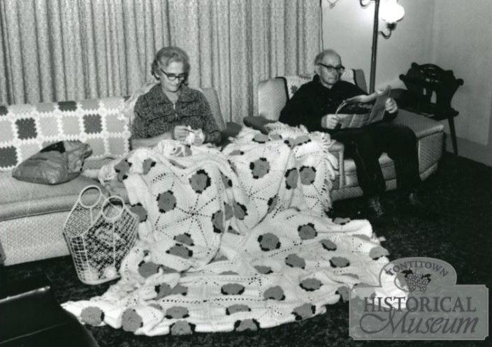 Woman sits on couch crocheting a large afghan; man sits in chair reading newspaper. In carpeted room, drapes in background, floor lamp to right of man. Knitting basket on floor left of woman.