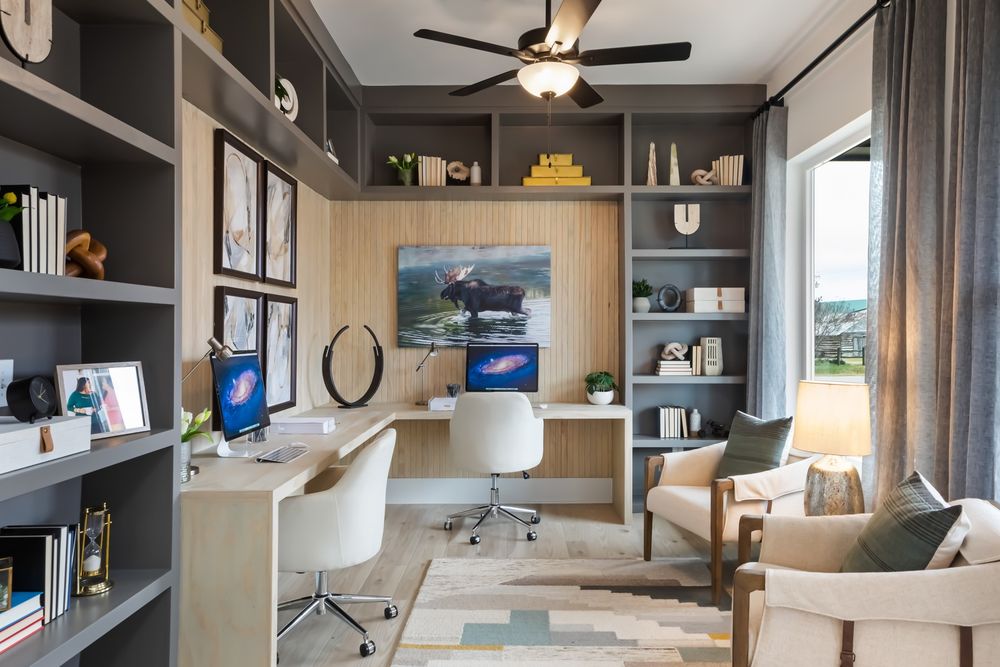 A modern home office by David Weekley Homes featuring a desk with dual monitors, a comfortable seating area, and built-in shelves with books and decorations. There is a large window providing natural light and a painting of a landscape hangs above a small cabinet.