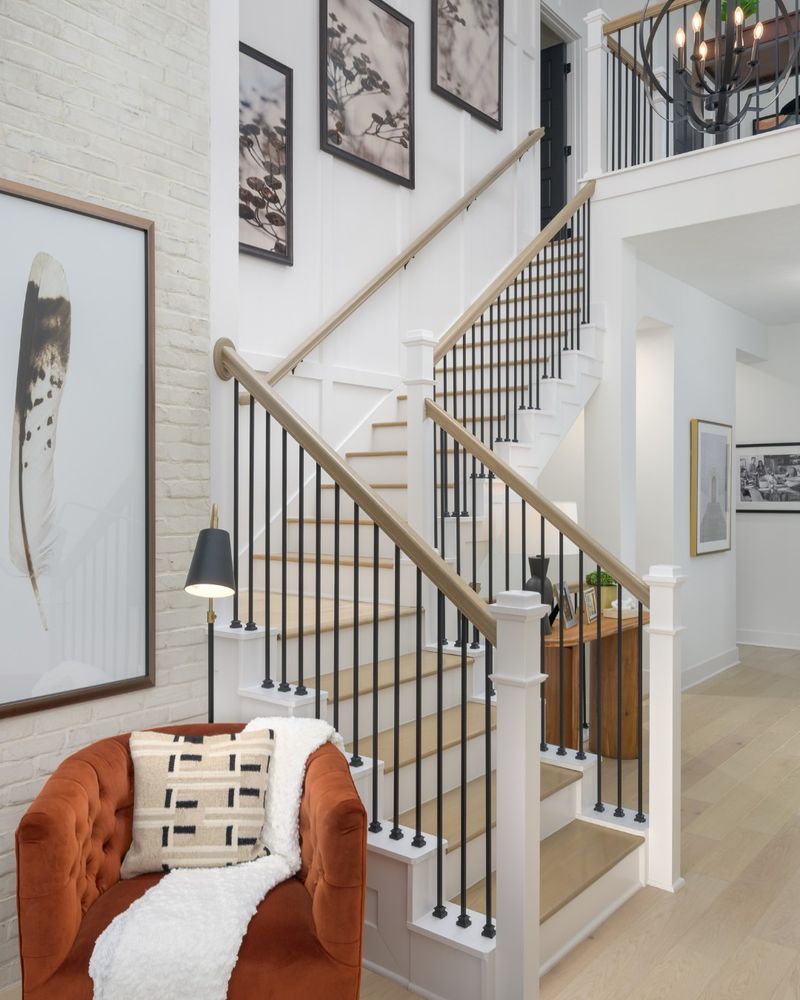 Interior view of a stylish staircase in a home by David Weekley Homes with white walls and wood flooring. Features include a cozy orange chair with a cushion, a white blanket, wall-mounted artwork, and a unique overhead chandelier.