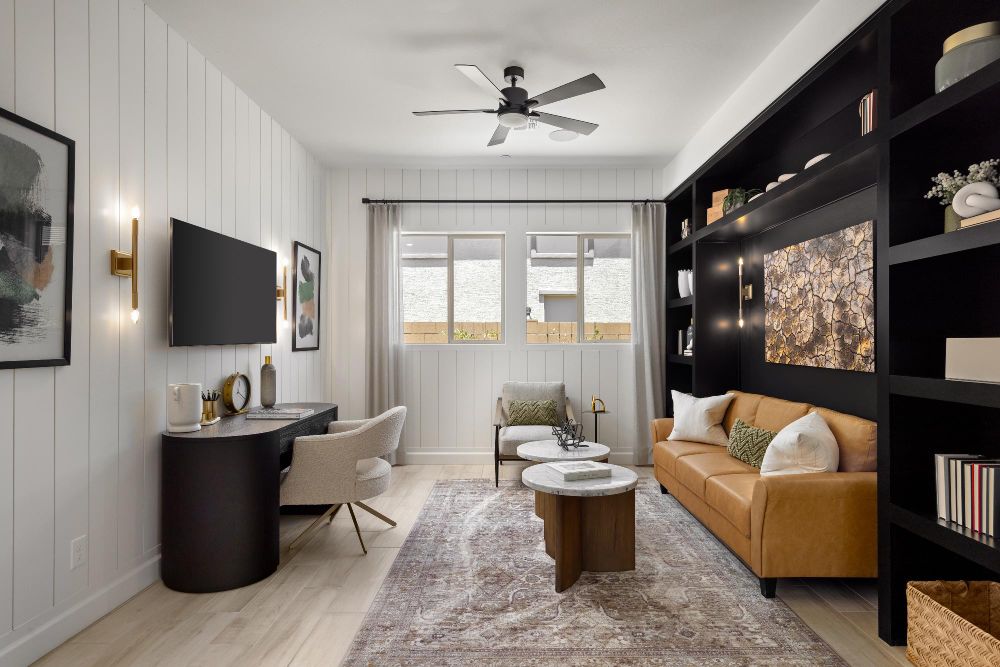 Modern home office by David Weekley Homes with white walls and dark shelving units. Includes a tan sofa, wooden coffee table, beige armchair, and a ceiling fan. Decorations include books, plants, and a wall-mounted TV.