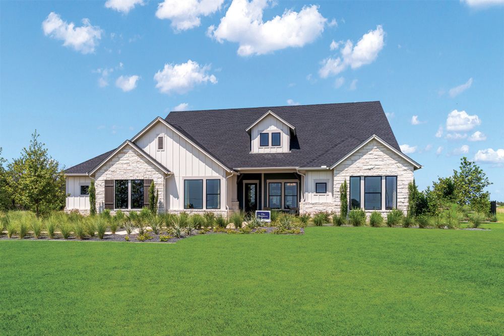 A single-story, modern farmhouse-style house with a large front yard, featuring stone accents and multiple windows under a clear blue sky. The Monterey model home from David Weekley Homes in Leander Estates is open daily for tours at 3085 Honeywood Drive, Leander, TX 78641.