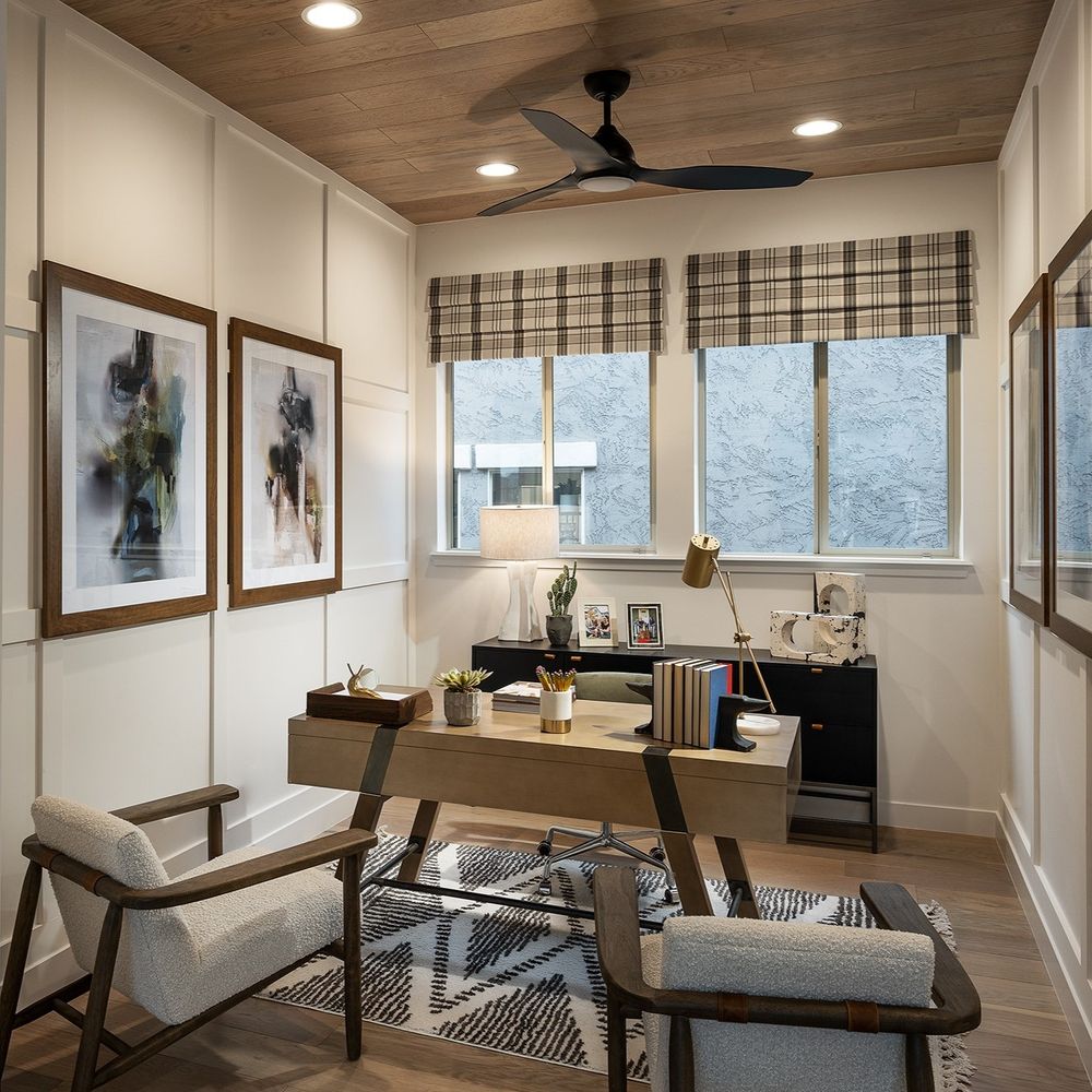Modern home office by David Weekley Homes with a desk, chair, bench, and wall-mounted pictures. The room features large windows with plaid blinds, a ceiling fan, and is decorated with plants and various office supplies.