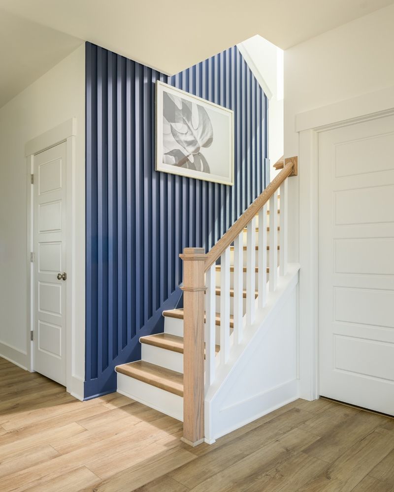 Interior view of a staircase by David Weekley Homes with wooden steps and white balusters, featuring blue and white striped wallpaper and a framed monochrome artwork on the wall. Two white doors are visible, one at the base and one at the top of the stairs.