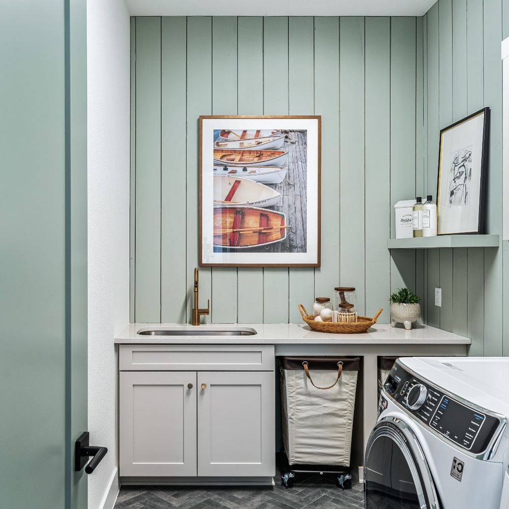 Interior of a laundry room by David Weekley Homes featuring a washer and dryer under a white countertop, gray cabinets, a framed picture of a boat on the wall, and a towel basket beneath the sink.