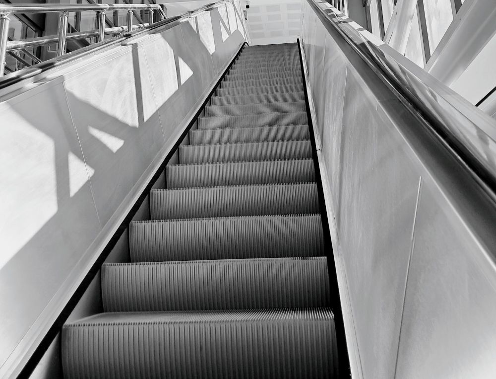 Picture of long escalator extending upwards, shot from low angle; sunlight from right casts geometric shadows on walls surrounding escalator