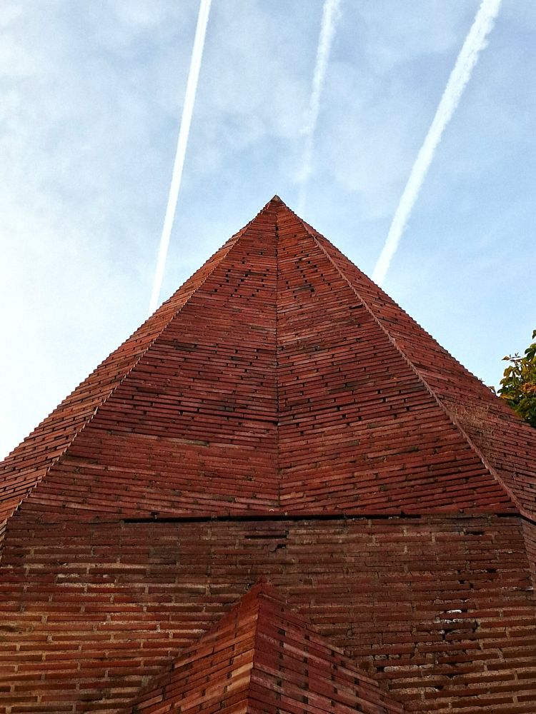 Picture of top of red brick building with pyramidal roof tapering to sharp point against partially cloudy blue sky; layered horizontal brickwork has textured surface on triangular faces with smaller pyramidal roof section emerging at bottom center; three white vertical contrails pass across sky behind structure on top of frame, sparse green tree leaves visible on right edge