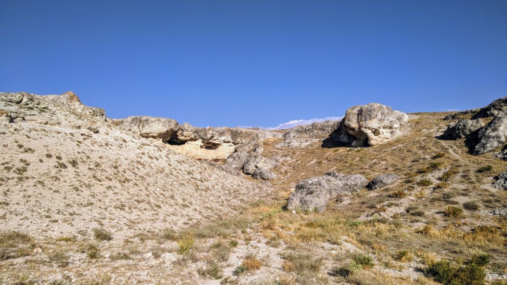 Picture of dry rocky terrain with scattered low-lying shrubs under clear blue sky; large rock formation with natural cave runs horizontally across middle of frame