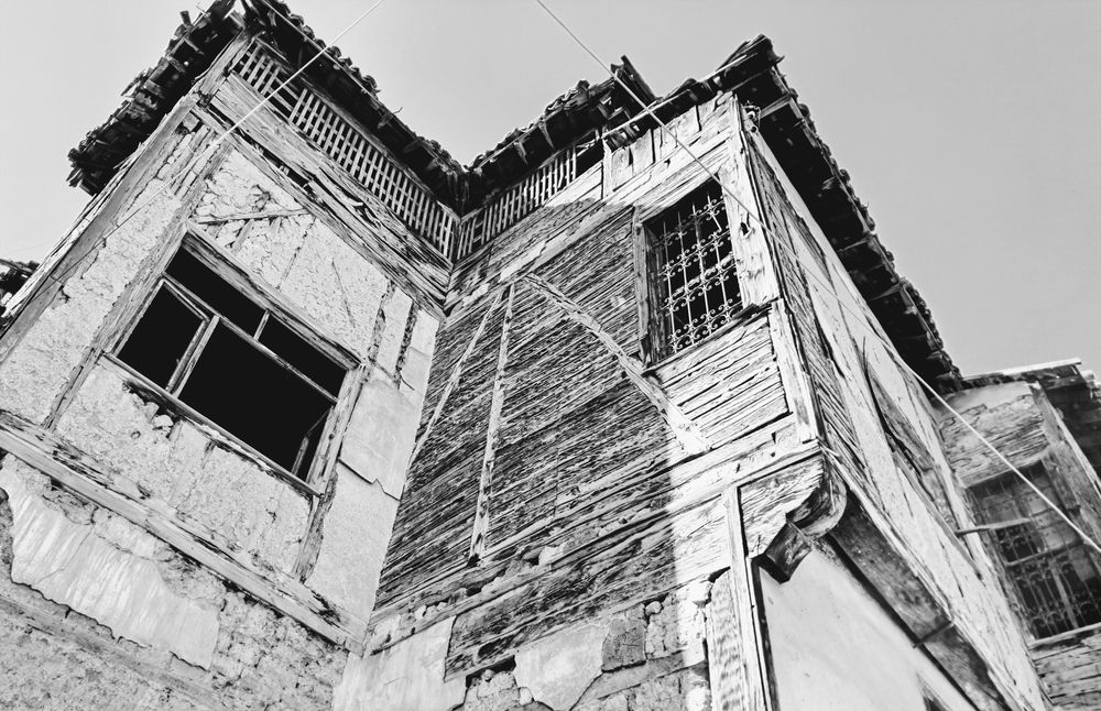 Picture of corner of derelict wooden and plaster building shot from low angle; building shows cracked mortar, exposed wooden beams, and boarded-up window with wrought iron grate, under clear bright sky