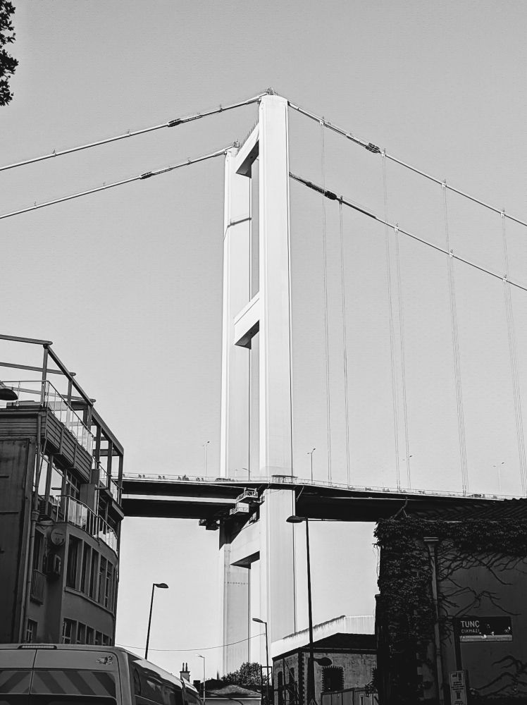 Monochrome picture of tall suspension bridge from ground level; white pylon with suspension cables in center and upper half of frame; low rise buildings, urban street, and street sign in lower half, all under clear blue sky