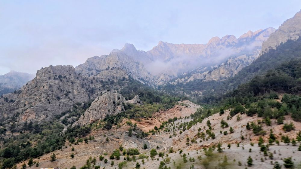 Picture of rugged mountain range with steep gray rocky peaks rising against hazy pale blue sky; sloping terrain with scattered green trees and shrubs on light brown rocky soil in foreground, misty upper slopes and valleys obscuring distant peaks in background; jagged rock formations on left side of frame, while right side shows more vegetation cover; early morning light casts soft shadows across landscape