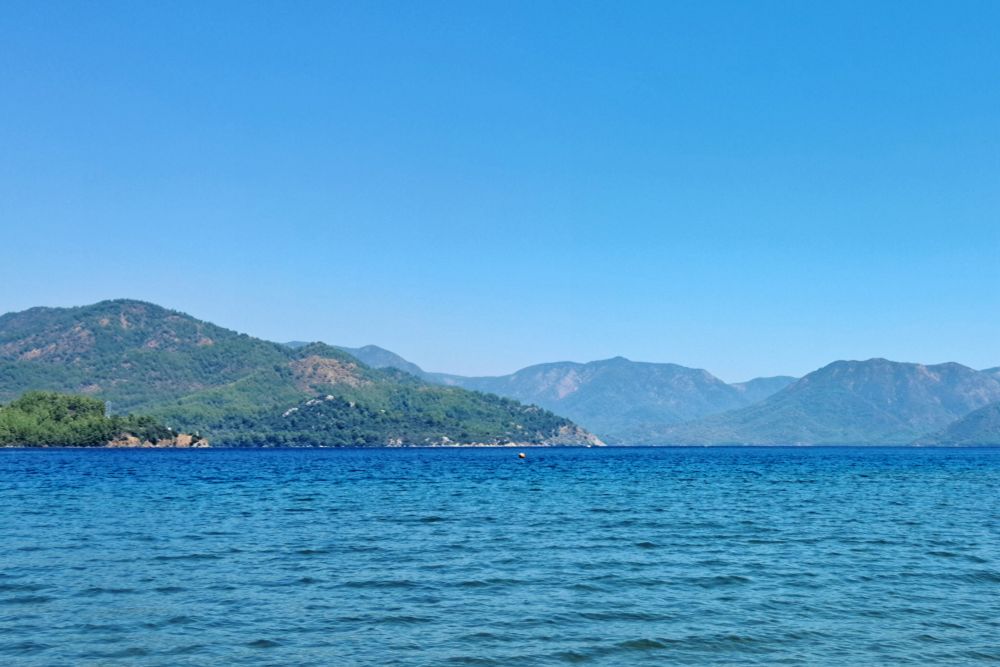 Picture of mountainous coastline with deep blue water under clear blue sky; water surface has rippled surface, green trees cover mountain foothills on left, rocky slopes on right and in distance; single small orange buoy floats on water near center horizon