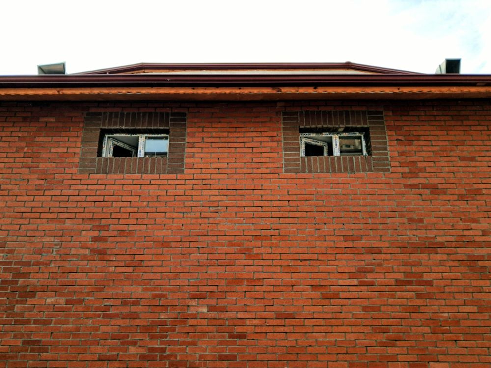 Picture of building exterior made out of red brick with two small rectangular windows with white frames and darker brick trim near top, brown eave overhanging brick wall, under cloudy white sky