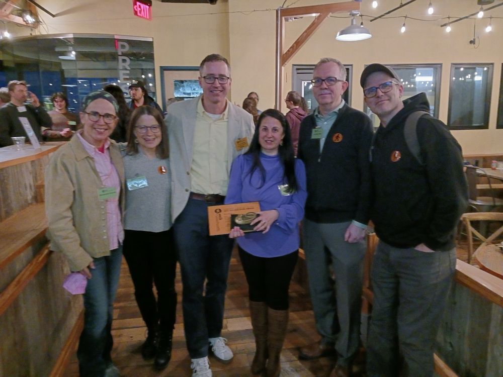 Picture of a group of people smiling. There are people in the background in a cafe setting