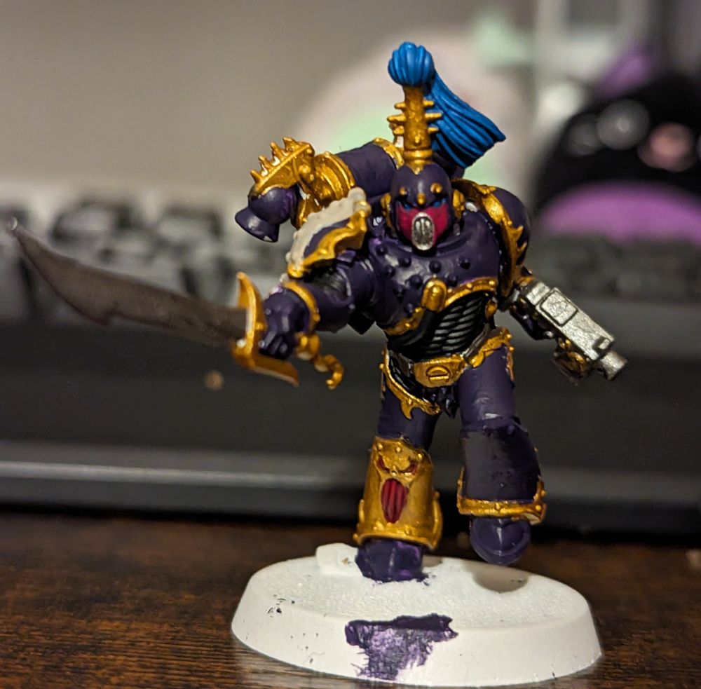 A Warhammer 40,000 miniature painted in purple with gold trim. The helmet has a pink mouthpiece and light blue top