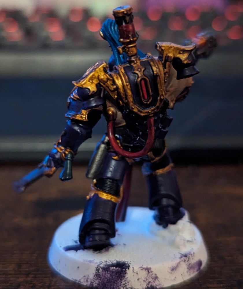 A Warhammer 40,000 miniature painted in purple and gold. Wielding a sword and handgun. He wears a white and purple helmet with blue and gold decoration