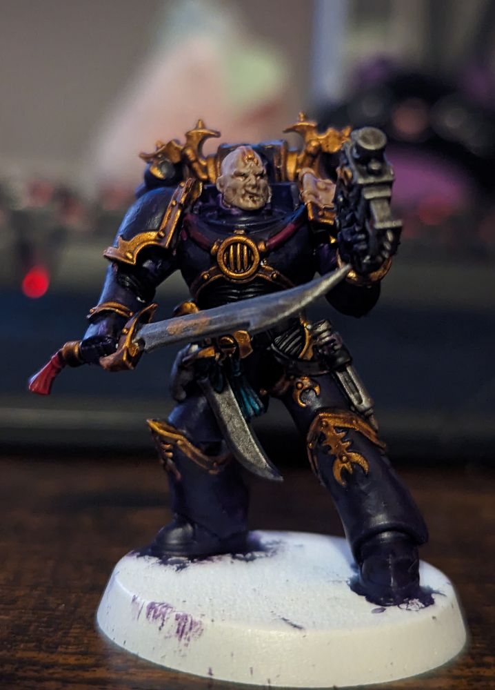 A Warhammer 40,000 miniature painted in purple with gold trim wielding a pistol and sword