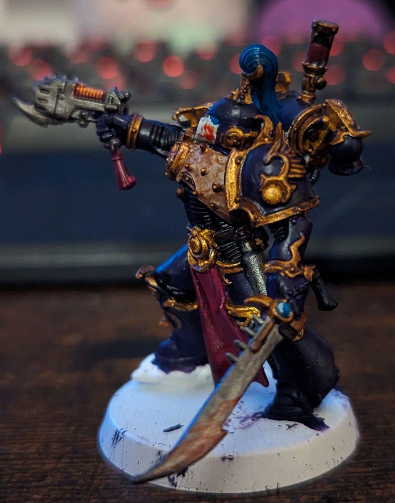 A Warhammer 40,000 miniature painted in purple and gold. Wielding a sword and handgun. He wears a white and purple helmet with blue and gold decoration