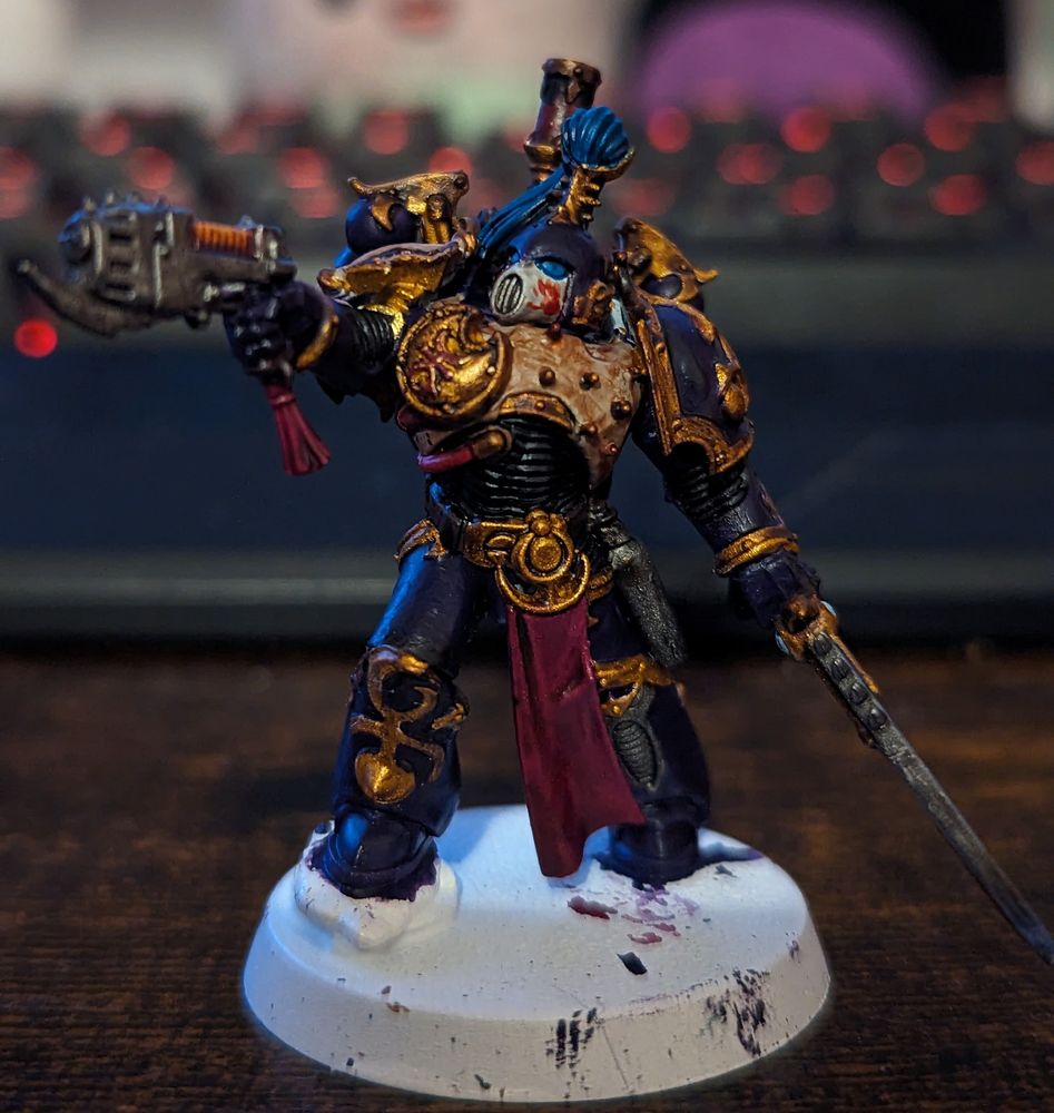 A Warhammer 40,000 miniature painted in purple and gold. Wielding a sword and handgun. He wears a white and purple helmet with blue and gold decoration