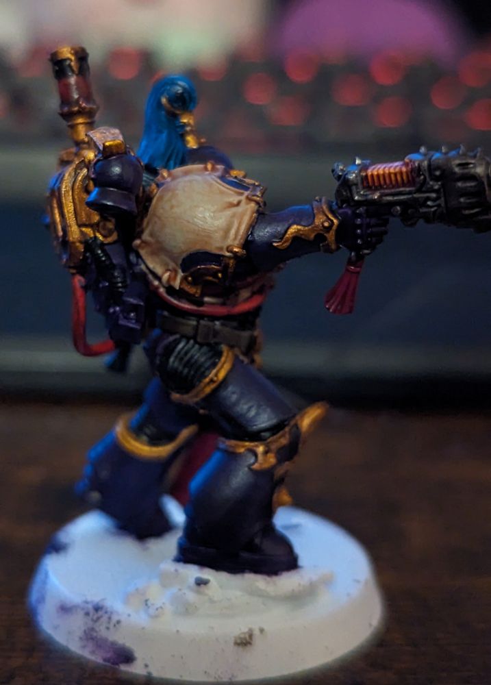 A Warhammer 40,000 miniature painted in purple and gold. Wielding a sword and handgun. He wears a white and purple helmet with blue and gold decoration