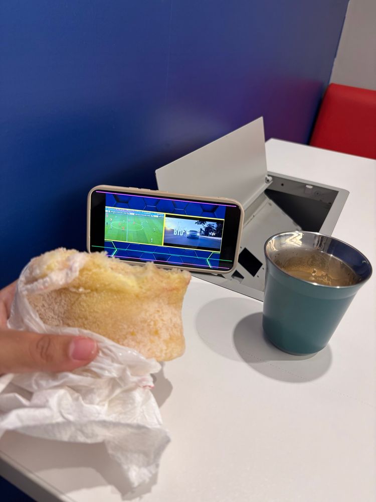 Mão sobre uma mesa branca segurando uma tapioca ao lado de um copo de café. Um celular apoiado em um suporte transmitindo o amistoso de futebol Coreia do Sul x Brasil