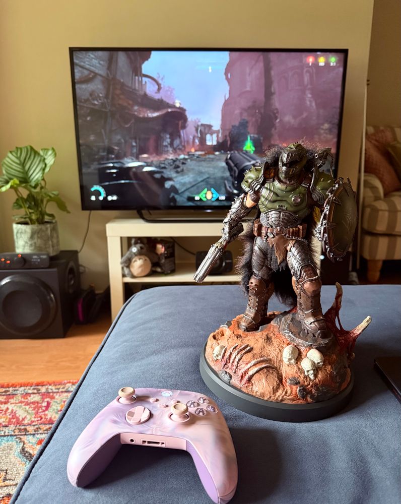 Statue of doom guy from doom the dark ages and a pink Xbox controller infront of a tv screen with doom the dark ages game running and a little green plant 