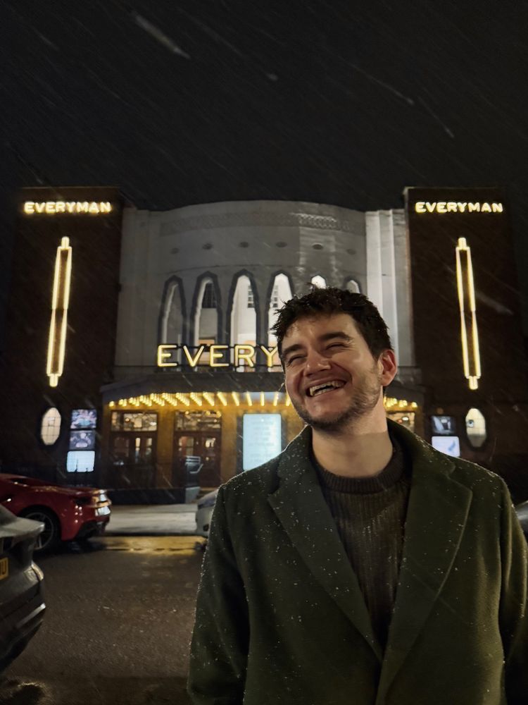 Picture of James in a green wool coat smiling over his right shoulder standing in front of the cinema 