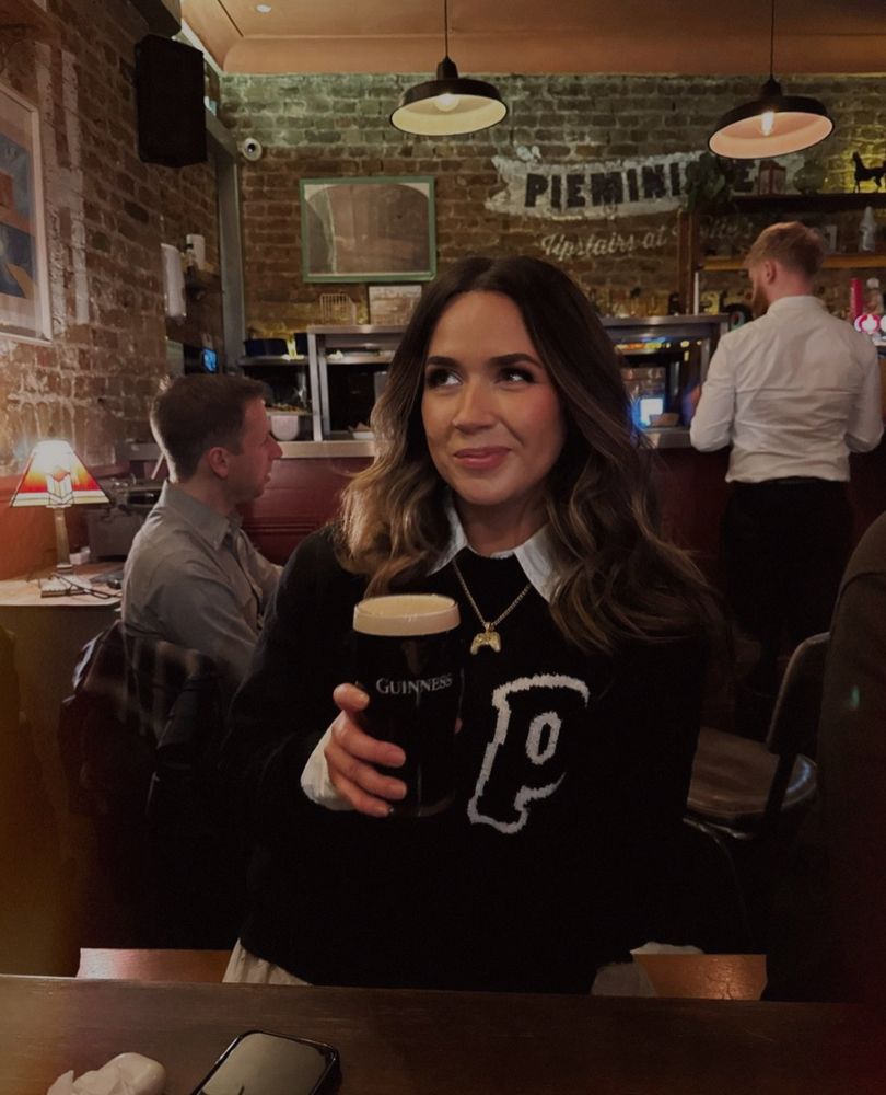 A picture of me at the pub smiling and holding a Guinness wearing a black jumper on a white collar shirt so it kind of looks like an outfit inspired by a Guinness 