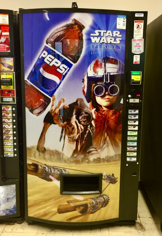 A Pepsi vending machine decorated with a large Star Wars: Episode I – The Phantom Menace promotional image. The design features young Anakin Skywalker wearing racing goggles and a helmet, standing beside Sebulba, his podracing rival. Below them, Anakin’s podracer speeds across the desert of Tatooine, leaving motion trails behind. A large tilted Pepsi bottle dominates the upper left corner.