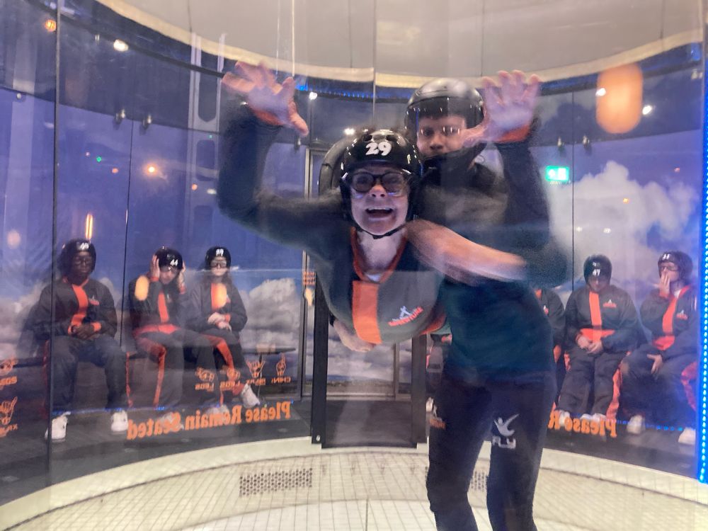 A woman in an indoor skydiving thingumie, being horizontally supported by the air currents created by industrial fans. a fella stood next to her is keeping her from spinning out of control.