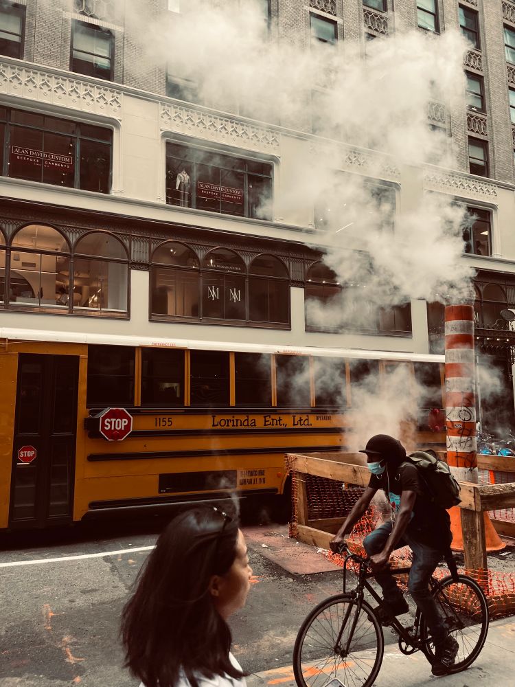 Steam rises out of an orange and white striped pipe connected to the street for some public works action while a yellow school bus passes in the background and a person on a bike and a pedestrian pass in the foreground