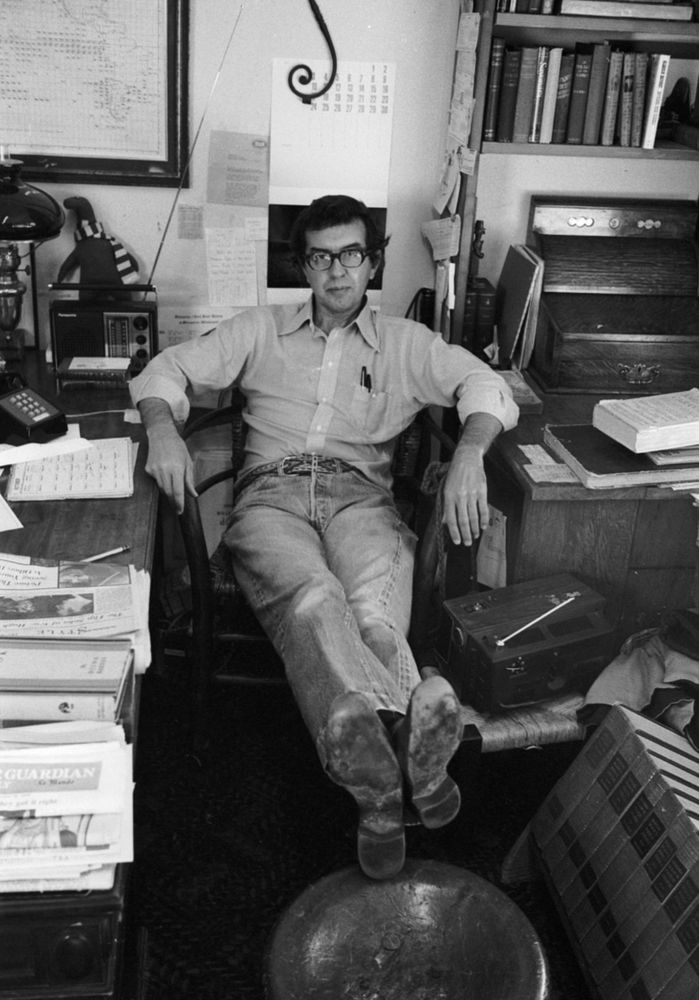 Larry McMurtry, seated, in jeans and cowboy boots, in the middle of a cluttered office