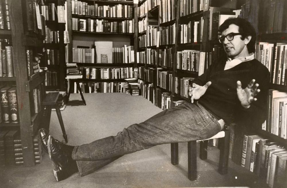 Larry McMurtry, seated, in a sweater, jeans, and cowboy boots, in a bookstore.