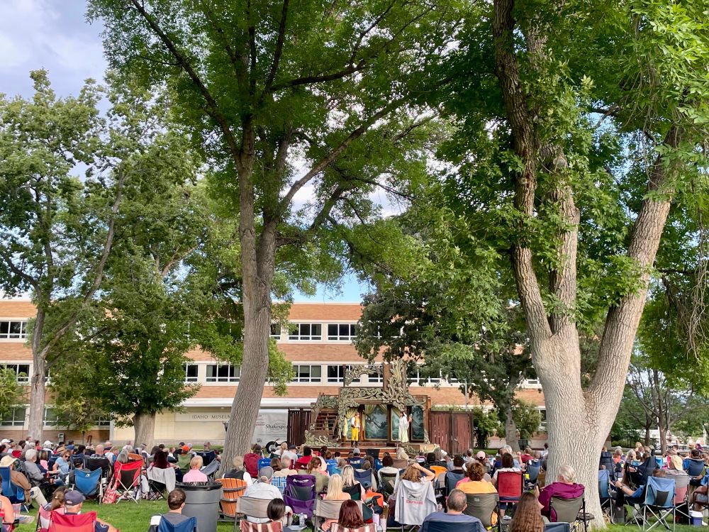 Outdoor performance of As You Like It, featuring a large audience, huge elm trees,  and a stage in front of a large brick building. 
