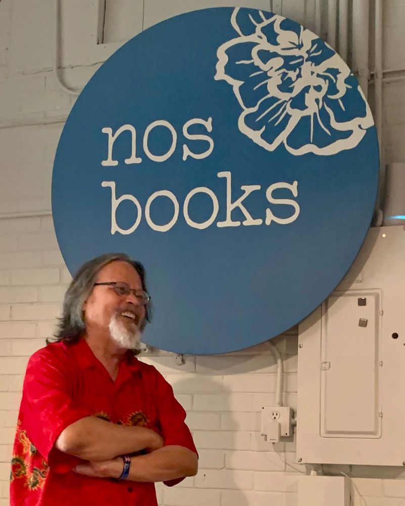 Iowa Poet Laureate Vince Gotera wears a red-dragon shirt. Behind him, the "nos books" logo is a white flower superimposed on a slate-blue circle.