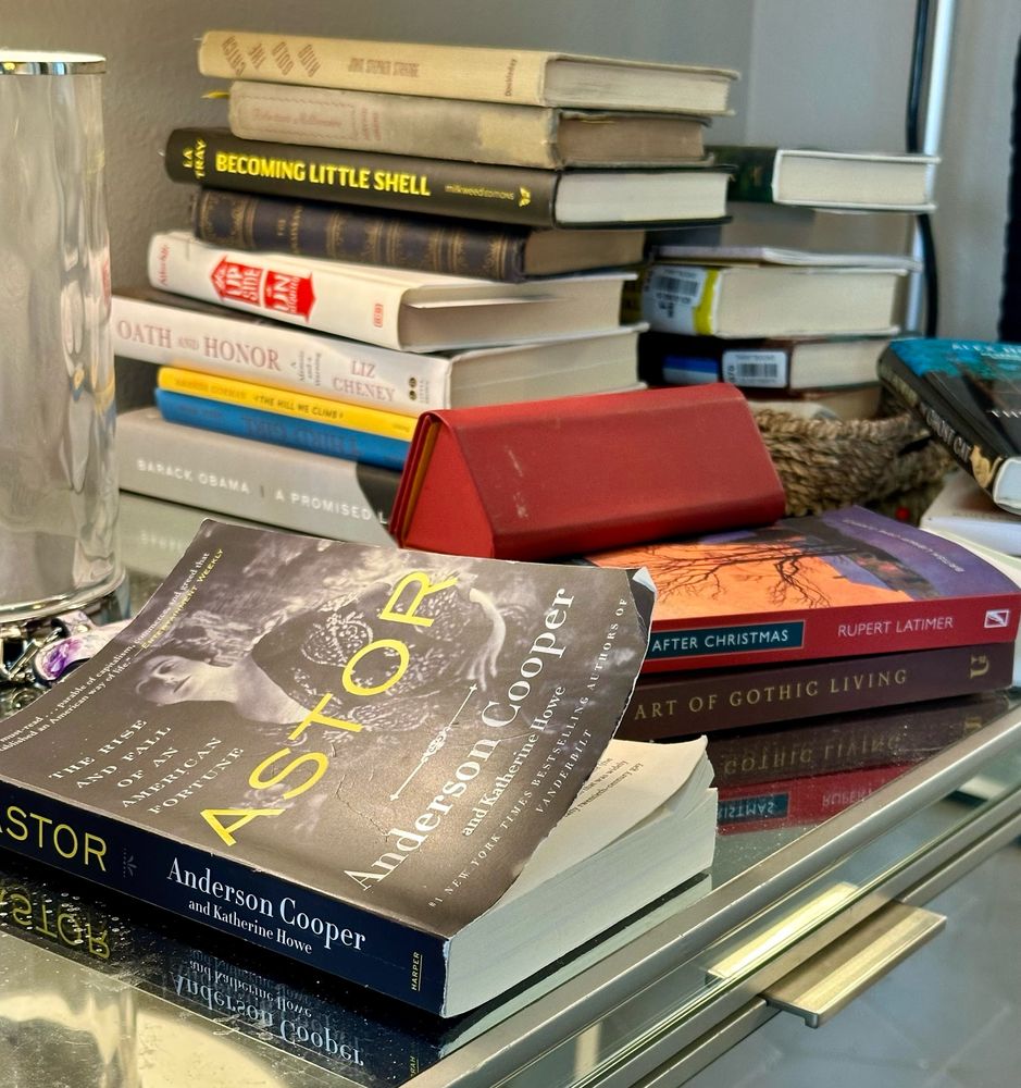 A disaster pile of various books cluttering a nightstand. Complete with water jug and roll of Tums. 
