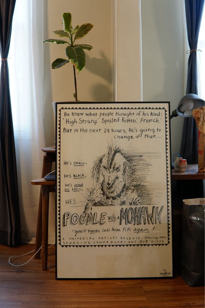 A photo that shows a framed poster sitting on the floor, leaning against a desk. The poster has a comic rendition of an angry looking poodle, with the text “he knew what people thought of his kind. “High strung” “spoiled rotten” “french” But in the next 24 hours he’s going to change all that. He’s small. He’s black. He’s mad as hell. He’s POODLE with a MOHAWK. “You’ll never call him FiFi again!” The artist is Lynda Barry
