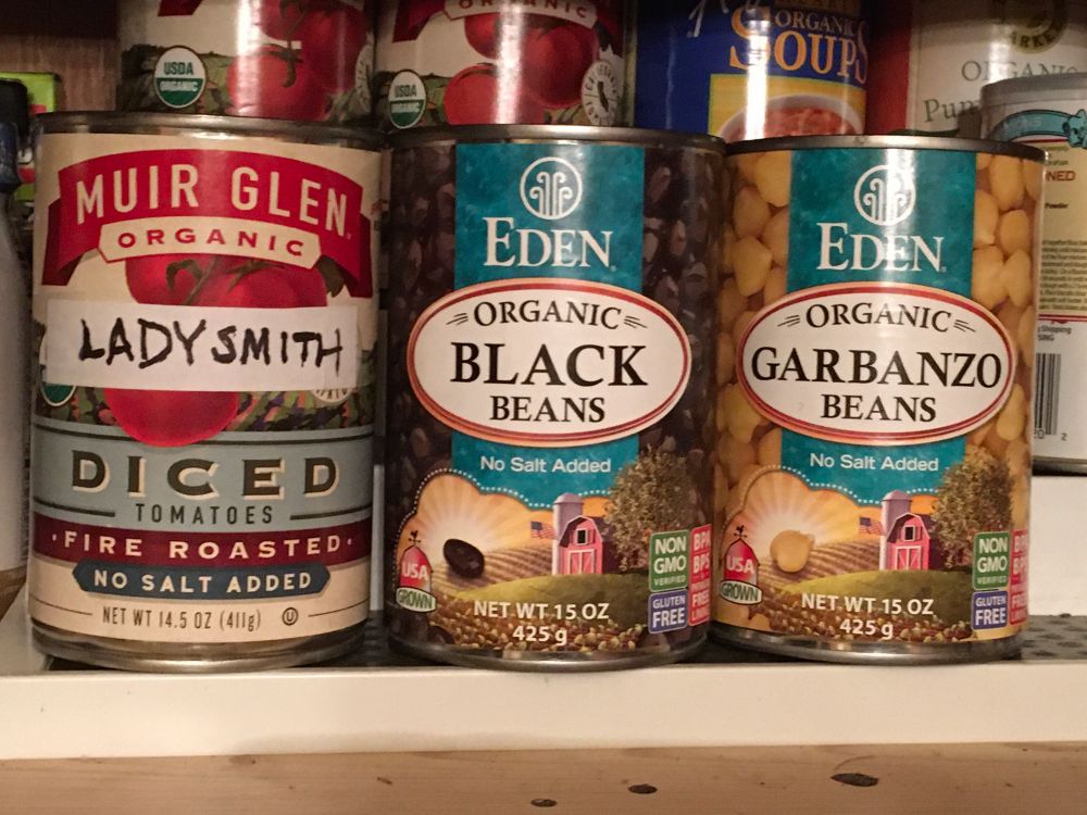 Three cans in a cupboard — tomatoes, Black Beans and Garbanzo Beans. The first has a handwritten note aligned to the next two labels. The handwritten note reads “Ladysmith” so that together it reads “Ladysmith Black Garbanzo,” similar to a well-known musical group.