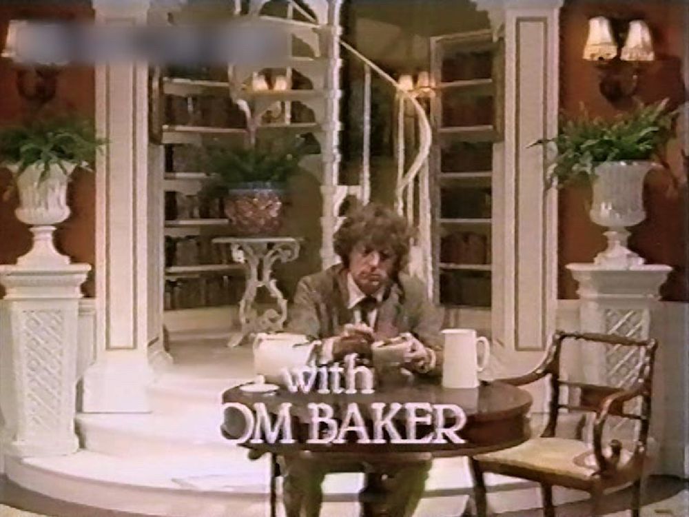 Tom Baker, in a brown grey scruffy looking suit & tousled brown mop of hair sits at a desk at the bottom of a winding staircase, surrounded by books in an a room that is otherwise painted almost all ‘James Bond baddie white’ 