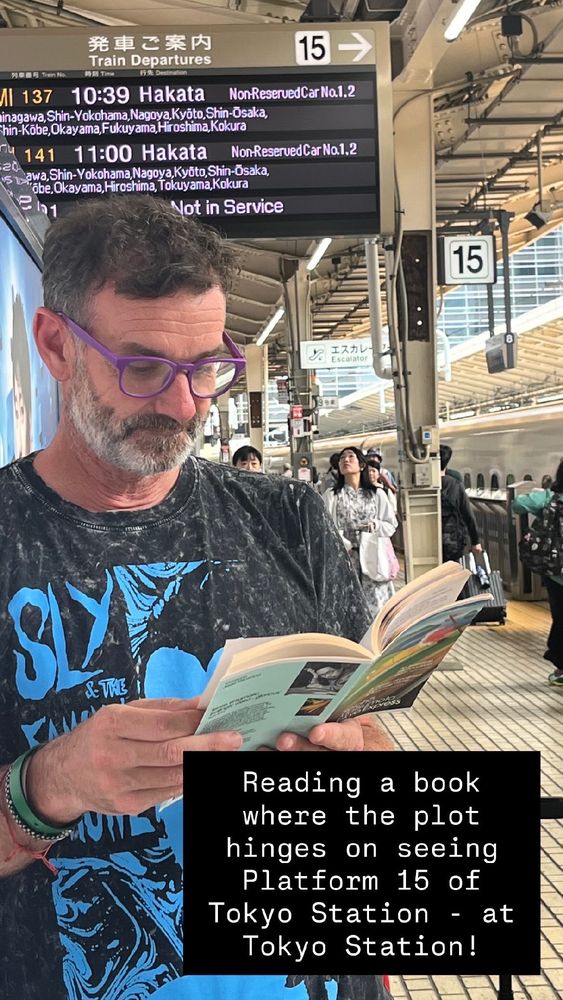 A dark-haired, entirely greying, light-skinned man wearing faintly ridiculous purple reading glasses reads a copy of Seicho Matsumoto’s Tokyo Express while waiting for a bullet train. He is wearing & Sly & the Family Drone tshirt which riffs off the snake priestess from the film  The Lair of the White Worm